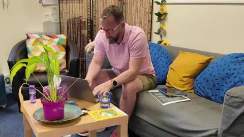 Andy Moreman is sitting on a sofa. He is working on a laptop which is placed on a coffee table.