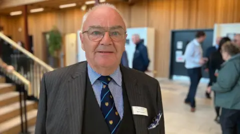 BBC/Seb Cheer A man wearing a suit and tie looks at the camera and smiles, in a modern building with people chatting in the background.