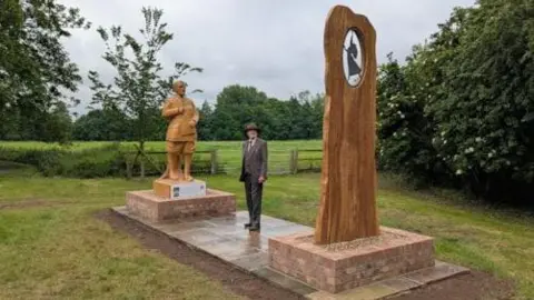 Wooden sculpture of JRR Tolkien depicted in his army uniform and holding a notebook, along with a second sculpture depicting the writer's wife Edith danncing. There is a man standing between the two structures dressed in a suit and hat.