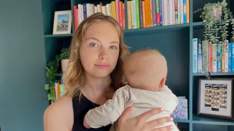BBC A woman with blonde hair and blue eyes looks at the camera while holding her baby, who faces away from the camera. In the background are blue shelves and books. 
