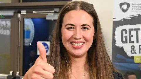 The Alliance Party's Sorcha Eastwood looks directly at the camera. She has long dark hair, has a big smile on her face and is giving a thumbs-up. 