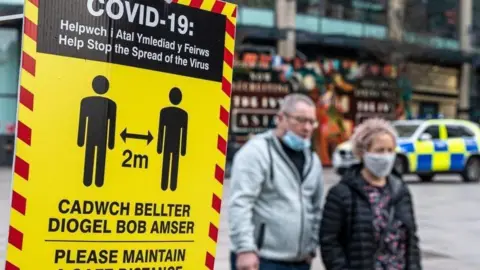 A yellow and red social distancing sign in the foreground to the left, with a couple walking past wearing face masks.
