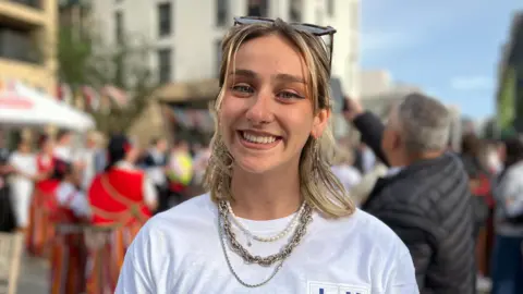 Hayley is wearing layered necklaces and a white “LUSO Project Luso” T-shirt smiling in a busy outdoor square, with a crowd, bunting, and people in traditional costumes behind.