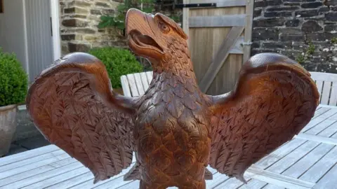 A wooden eagle sculpture on a table in what looks like a garden.