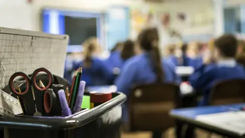 Image shows a classroom scene. Pupils are sat facing a blackboard. In focus at the front of shot is a pot of classroom equipment including scissors and pens.