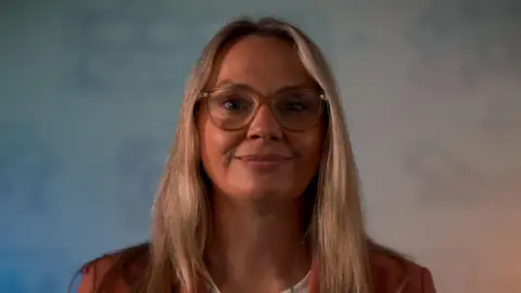 A photograph of a woman wearing glasses looking directly at the camera. She has blonde hair. 