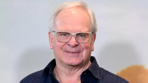 Getty Images Headshot of Cornwell. He wears a navy shirt, has short white hair, and thick grey framed glasses. 