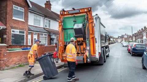 A bin lorry stops on a residential street with two bin men loading black bins into the back of the lorry before returning them to the kerbside.
