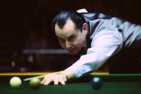 Reuters Ray Reardon, pictured in his playing days. He is leaning over the snooker table, getting ready to hit the white ball. He is wearing a white shirt and waistcoat favoured by nearly all snooker players. His hair is very dark and slicked back into a "widow's peak"