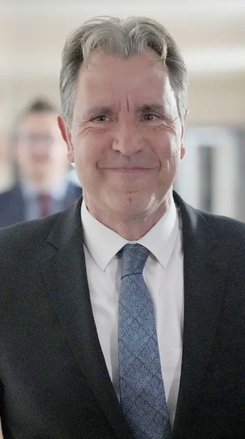 Dan Norris is walking down a corridor wearing a suit and smiling.