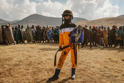 FREDRIK LERNERYD / AFP / GETTY IMAGES A  young jockey poses for a portrait. People stand in the background behind him, and the mountains rise in the distance.