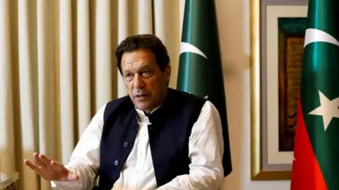 Former Pakistani Prime Minister Imran Khan speaks with Reuters during an interview, in Lahore, Pakistan March 17, 2023. He can be seen seated with his hand outstretched and speaking. Two Pakistani flags are behind him