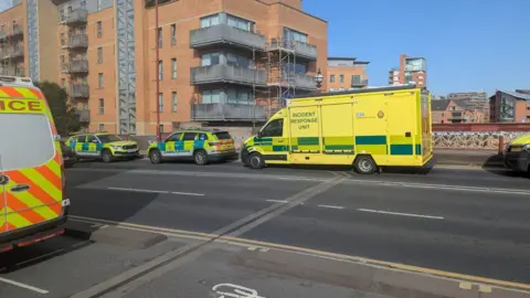 BBC/Steve Jones Emergency vehicles are parked along a city street outside modern brick apartment buildings. A large yellow vehicle labelled “Incident Response Unit” is positioned beside two police cars with high-visibility markings. Part of an ambulance is visible in the foreground, and scaffolding can be seen on one of the nearby buildings.