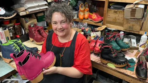Jackie holds a pair of bright pink clog boots, behind her are blue green red orange leather upper clogs 
