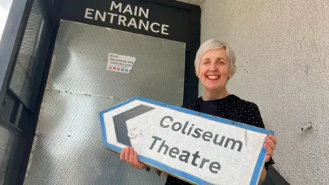 Julie Hesmondhalgh holding up a road sign saying "Coliseum Theatre" in front of the venue's boarded-up main entrance