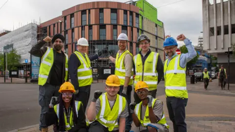 City of Wolverhampton Council A group of people wearing helmets and hi-vis jackets stand in front of the new college campus. The building has terracotta coloured cladding. Some protective covering and scaffolding can be seen on the exterior. Street signs and boards can be seen in front of the building.