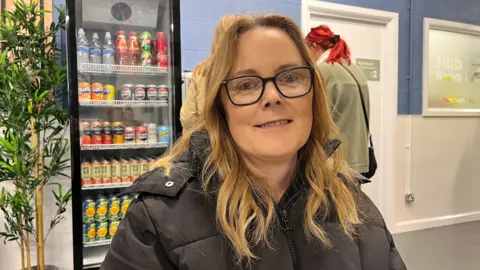 A woman with long ginger hair smiles as she sits at a table in the community cafe with a vending machine full of colourful cans of drink in the background. She is wearing a black coat and black-framed glasses. A green plant stands to the left.