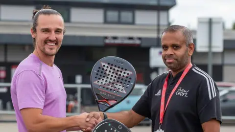 Callum Slater shakes hand with another man. They are both holding black padel rackets in their other hands. Callum wears purple T-shirt and has dark hair that has been tied up behind his head. The other man wears a black T-shirt with a red lanyard around his neck. He has short black and grey hair. Both men have short dark beards.