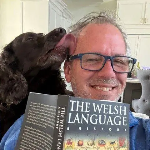 Jeff Narvil Jeff taking a selfie holding a book with the title 'the welsh language - a history' while a brown spaniel licks his ear