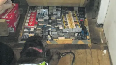 LDRS A black sniffer dog in front of a hidden compartment under some floor boards filled with contraband cigarerettes