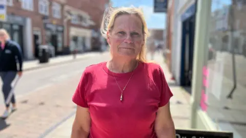 A lady with blonde hair and a pink top. She's standing on a high street with people shopping in the background. It is a sunny day.