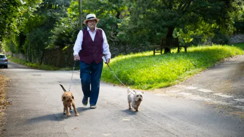 Robin Shuckburgh on a country road walking his dogs, Gizmo and Widget. They are walking by a green space. It is a sunny day.