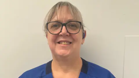 University Hospitals of Northamptonshire A woman with brown hair and glasses, wearing royal blue hospital scrubs standing in front of a white wall. 