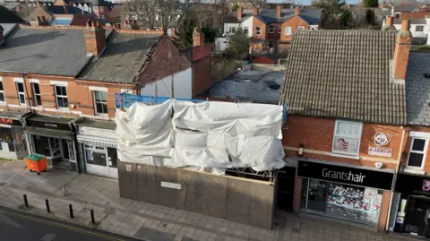 An aerial shot of a building which does not have a roof or upper floors surrounded by white sheeting and boarding