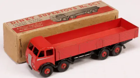 Vectis Auctions A long red toy truck is placed beside its cardboard box. The vehicle has four visible black wheels and holes for windows around the cab at front. The wagon behind it is open. The box has a red design on it reading: "Dinky Supertoys no. 501. Foden Diesel 8-Wheel Wagon."