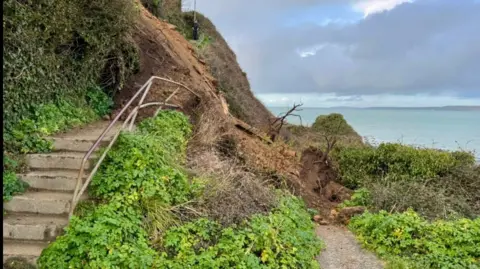 Newquay coastguards The picture shows a coastal footpath that has been hit by a landslide. A section of the hillside has collapsed, sending soil, rocks and vegetation down across the path. The metal handrail along the steps is bent and partly buried by the slip. Green undergrowth surrounds the area, and the sea is visible in the background under a cloudy sky. 