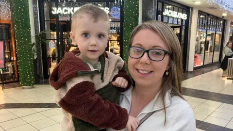 A woman with blonde hair, a white top and glasses is holding a young child inside a shopping centre. The child has blonde hair and is wearing a multi coloured top.