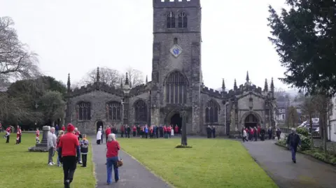 Danny Lawson/PA Wire A view of Kendal Parish Church - built in the gothic style in white limestone with a central tower. People are walking across a the grassed area in front of it and there is also a gathering at the entrance. Many are wearing red as requested by the family of Poppy Atkinson whose funeral it is.