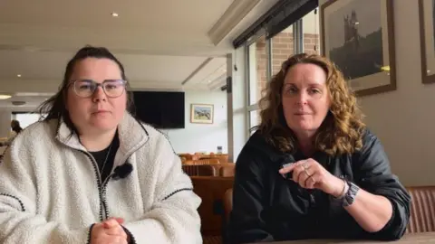 Two women sitting at a table in a cafe with white walls and pictures of racehorses on the walls. The woman on the left has black hair, tied back, and wears a white fleece. The woman on the right has curly brown hair and wears a black jacket. 