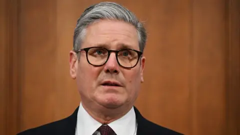 Getty Images A man with grey hair and black glasses photographed at a press conference. He is wearing a black suit and white shirt and is visible from the shoulders up. Behind him is a wood-panelled wall.