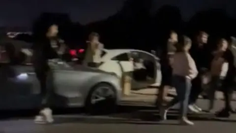 Supplied A night-time roadside scene showing several people gathered beside a parked car with its door open. Other vehicles are nearby. The image is taken from phone camera footage, with dark surroundings and limited lighting from vehicles.