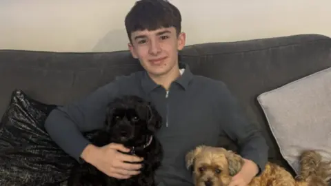 South Central Ambulance Service A teenager sat on a sofa smiling while cuddling a black and a brown dog 