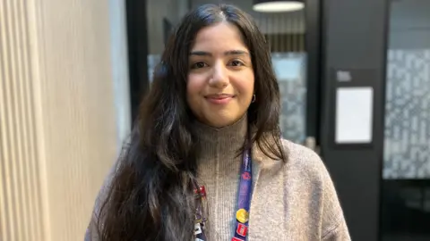 A woman with long dark hair and a grey top, with a purple lanyard around her neck, smiles as she stands in a room.