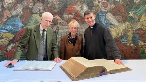 Kerry Eaton (middle) and the Reverend James Harvey (right) standing with another man next to a large ancient large bible opened on a table. They are smiling while behind them is a giant religious artwork. The man on the left wears a green jacket and tie with a grey woollen waistcoat. He has white, receding hair and glasses perched halfway down his nose. Kerry Eaton wears a brown jacket and a dark scarf with white polka dots. She has short blond hair. The reverend has his dog collar and a dark grey fleece jumper. He has short dark hair with a parting on his left.