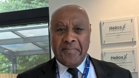 A man wearing a black jacket and tie, white shirt and blue lanyard is looking at the camera. He is standing near a wall and a window, with a wooden fence behind it, is visible on the left of the photo.