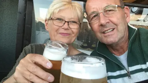 Family photo Ross and his wife holding up pints of beer smiling and looking towards the camera. Mrs Leslie is wearing a brown top and Mr Leslie a green and white striped top.