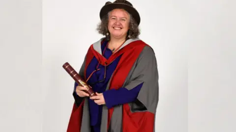 University of York A person is standing in a studio setting wearing academic graduation regalia, including a red and grey gown and a dark hat. The person is holding a rolled diploma or certificate tied with decorative bands and is facing the camera. The background is plain and light-coloured, placing focus on the formal attire and the certificate being displayed.