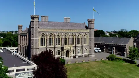 Victoria College in Jersey. The main building is made from grey stone and has large church-like windows. The building has turrets on the corners. A large green lawn is in front of the building. It is a sunny day.