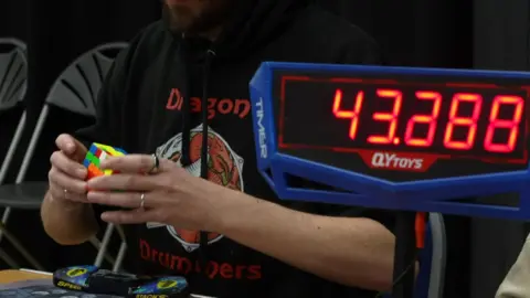 A person solving a Rubik's cube. There is a timer in front of it. The person is using two hands to solve the cube and is wearing a black jumper.