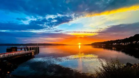 Tommy Bamford An orange sunrise over a calm body of water. The sun is just rising above the horizon and the sky is a mixture of blue, grey and orange. The water is still and calm and is reflecting parts of the sky. A metal and stone pier stretches out into the water on the right hand side.