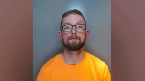 Durham Police A mugshot of a man with short, brown hair and glasses wearing a bright yellow t-shirt.