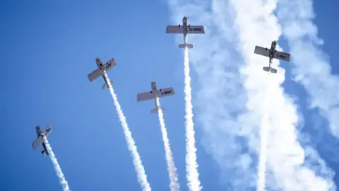 Five small aerobatic aircraft flying overhead against a bright blue sky looking almost straight up from below, so the undersides of the aircraft are visible. Each aircraft is producing a thick white smoke trail that extends downward through the image. The smoke trails are nearly vertical and vary slightly in angle, creating a fan-like pattern across the sky. 