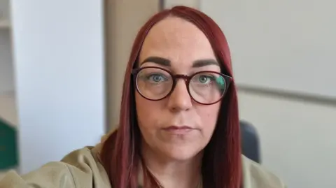 A woman with long red hair and red glasses looks into the camera. She is wearing a beige top. Light-coloured walls are in the background.