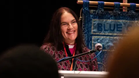 A female priest in pink, black and yellow robes, smiling at a lectern behind a microphone. She has long black hair and is standing next to a blue fabric hanging banner.
