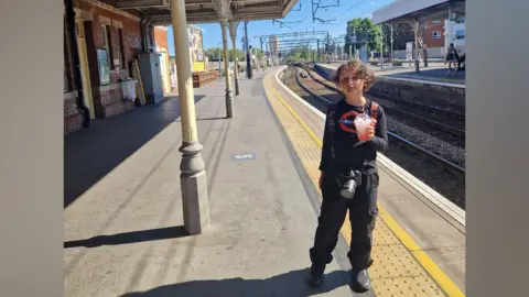A picture of George waiting at a train station on the day of the challenge. George is wearing all black, and his top has a TfL roundel on it. He has longish brown hair and is holding a cold drink. He also has a bag on his back and camera around his neck.