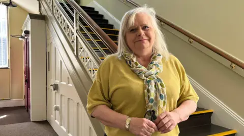 Anna Pugh stands by the stairs inside a building. She is wearing a yellow jumper and coordinated patterned scarf.